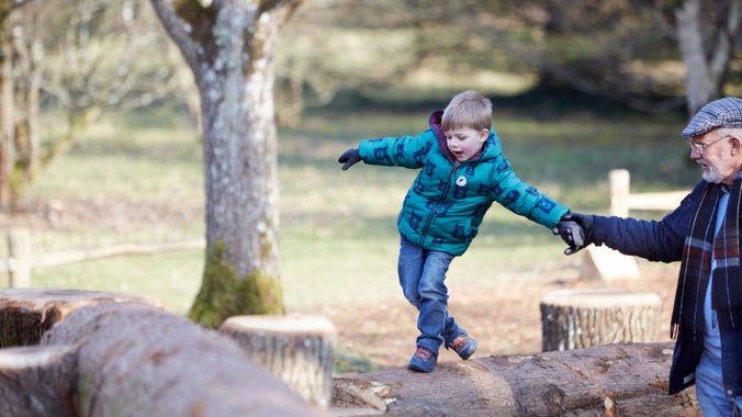 A little boy in winter clothes balances along a big log, helped by a older man